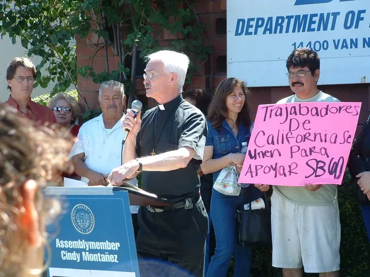 A priest speaking at a podium beside workers holding signs in support of SB 60
